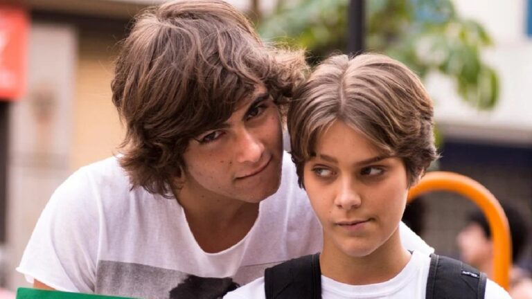 Dois jovens aparecem em primeiro plano em uma cena ao ar livre. O rapaz, com cabelo castanho médio e levemente ondulado, inclina-se por trás da garota e olha para ela com expressão suave. A jovem, de cabelo curto e castanho claro, usa mochila nas costas e desvia o olhar para o lado, com semblante pensativo. Ao fundo, há elementos urbanos desfocados, como árvores e construções, sugerindo ambiente de rua ou pátio escolar.