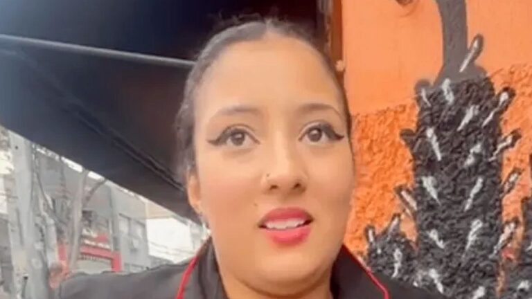 Texto Alternativo disse: Mulher aparece em primeiro plano gravando vídeo em ambiente externo urbano. Ela usa delineado marcante e batom vermelho, com expressão atenta, enquanto ao fundo há fachada laranja, estrutura metálica e elementos decorativos escuros, sugerindo registro informal para redes sociais.