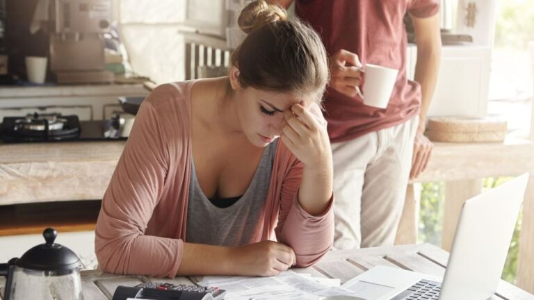 Mulher sentada à mesa com expressão de preocupação analisa contas e faturas ao lado de um notebook e calculadora, enquanto homem segura uma xícara