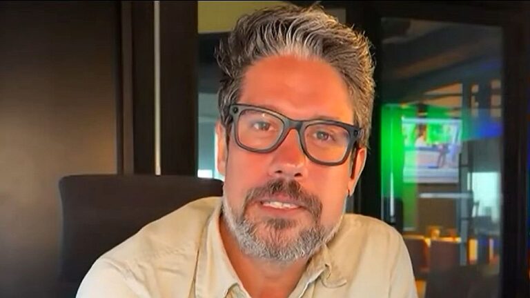 Homem de meia-idade com cabelo grisalho e barba bem aparada olha diretamente para a câmera. Ele usa óculos de armação escura e camisa clara. Está em um ambiente interno que parece um estúdio ou escritório, com iluminação quente e uma tela ao fundo exibindo cores verdes e imagens desfocadas. A expressão dele é séria e atenta, como se estivesse falando ou gravando um vídeo.