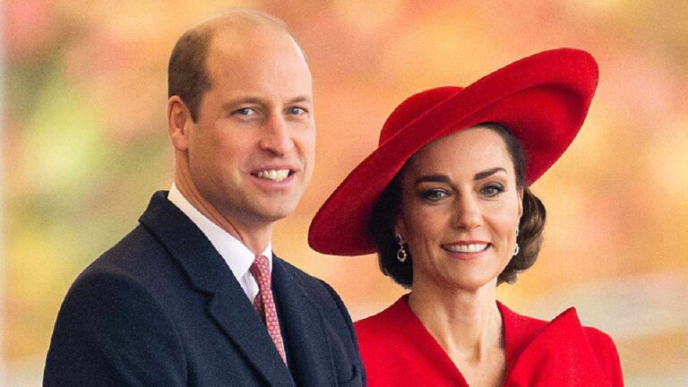Príncipe William e Kate Middleton sorrindo lado a lado em evento oficial usando trajes formais com destaque para vestido e chapéu vermelhos