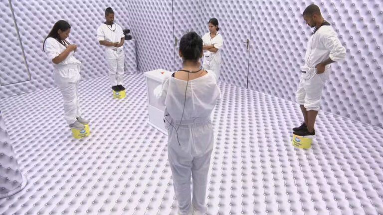 Cinco participantes do BBB 26 vestem macacões brancos e estão posicionados sobre bases amarelas em um quarto totalmente revestido por estofado branco