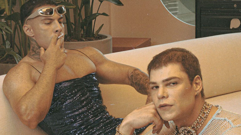 Rafa Marttinz fuma de óculos escuros e vestido de brilho ao lado de Fábio Marques o Tadalafellas que usa blusa de rede e correntes em sofá bege