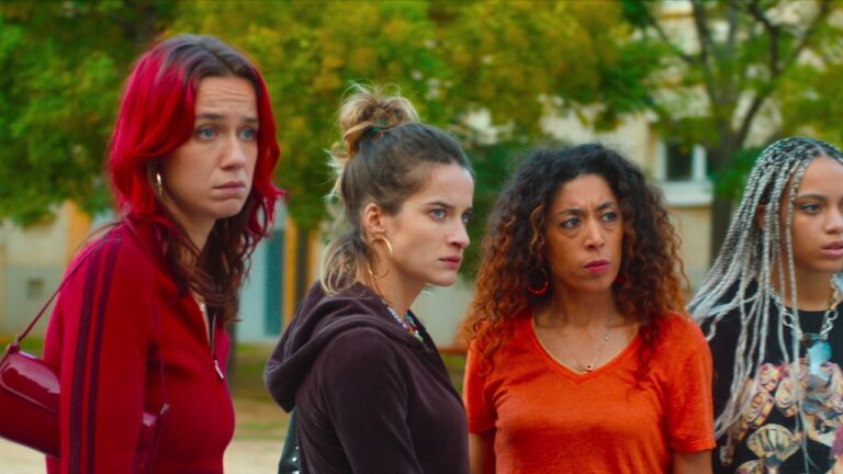 Quatro mulheres com expressões tensas observam algo fora de quadro em área residencial ao ar livre em cena de filme ou série dramática