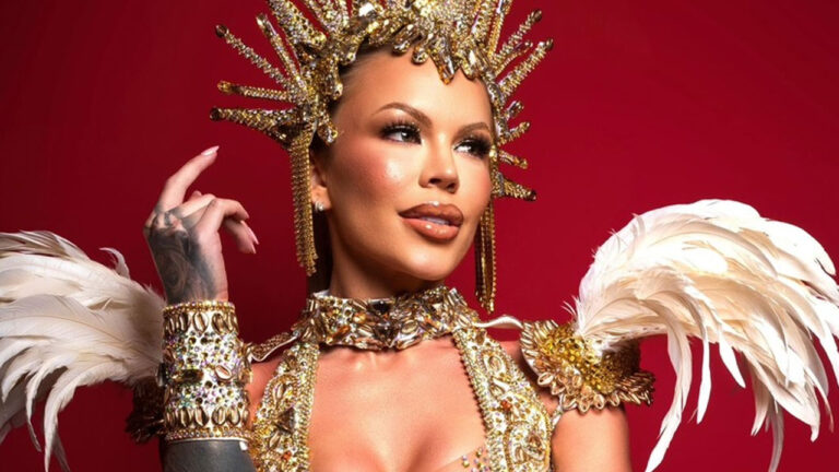 Ravena Hanniely posa com fantasia de luxo dourada e cravejada de cristais, usa coroa majestosa, ombreiras de penas brancas e fundo vermelho vibrante