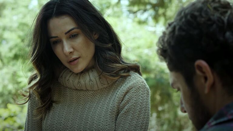 Mulher de cabelos longos e castanhos usa suéter bege de gola alta enquanto conversa com homem em área arborizada ao ar livre