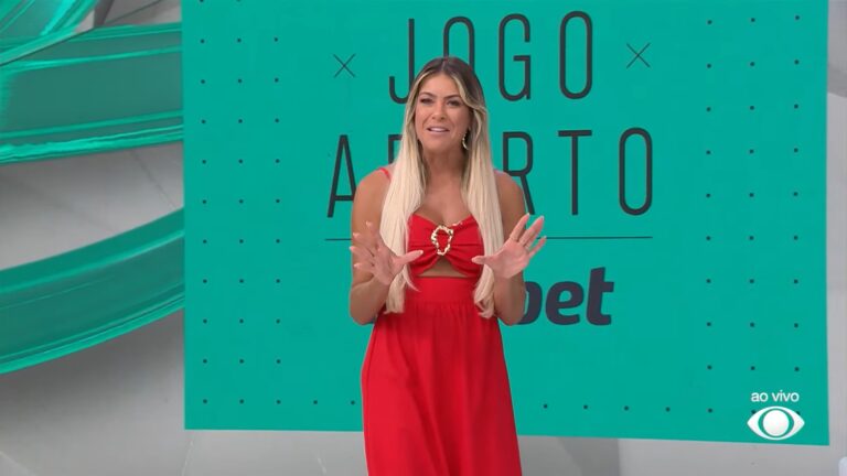 Renata Fan, uma mulher de cabelos longos e loiros, apresenta programa ao vivo em estúdio, vestindo vestido vermelho com detalhe dourado na parte frontal, gesticulando com as mãos; ao fundo, painel verde com o título Jogo Aberto.