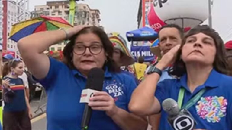 Duas repórteres usam camisas azuis e seguram microfones durante transmissão ao vivo em evento de rua, com público e bandeiras coloridas ao fundo.