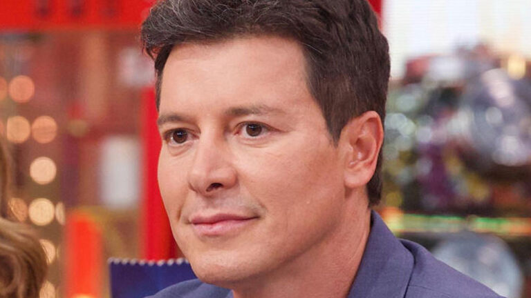 Apresentador Rodrigo Faro em close frontal com expressão serena veste blazer azul sob luz de estúdio televisivo com fundo levemente desfocado em vermelho