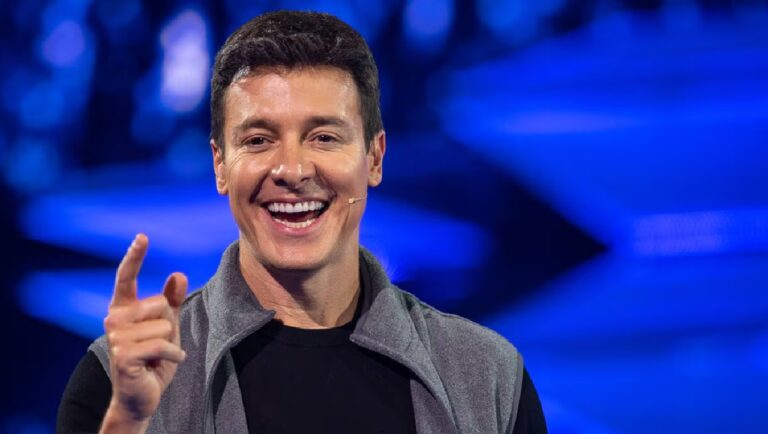 Rodrigo Faro sorri e aponta para a frente durante apresentação em estúdio de TV, usando camiseta preta e colete cinza, com microfone de lapela e fundo iluminado em tons de azul.