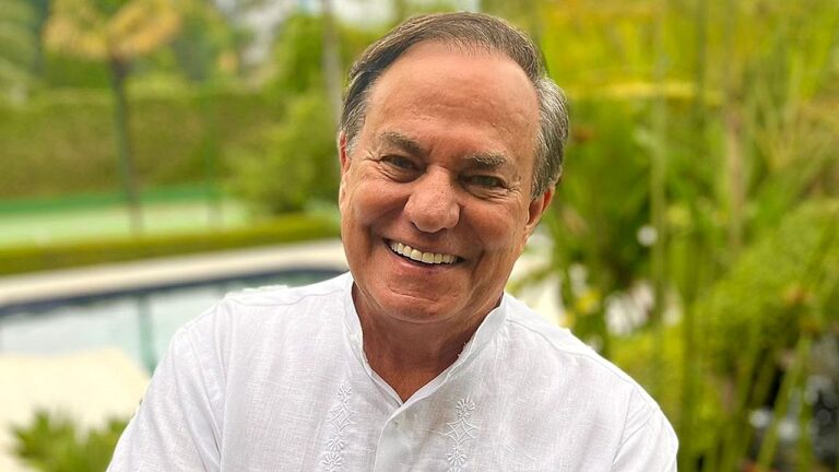 Homem de meia-idade sorri para a câmera em ambiente externo, usando camisa branca de tecido leve. Ao fundo, aparecem piscina e jardim com vegetação verde, sugerindo área residencial ou de lazer. A imagem transmite clima descontraído e ensolarado.