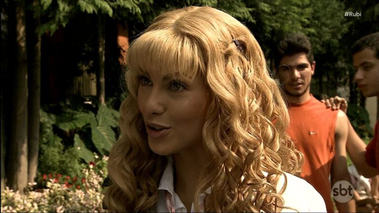 Rubi, usando uma peruca loira de cabelos longos e cacheados, conversa em um jardim arborizado; ela usa camisa clara e olha à frente com expressão firme. Ao fundo, dois jovens, um deles com camiseta laranja, observam a cena entre árvores e canteiros de flores