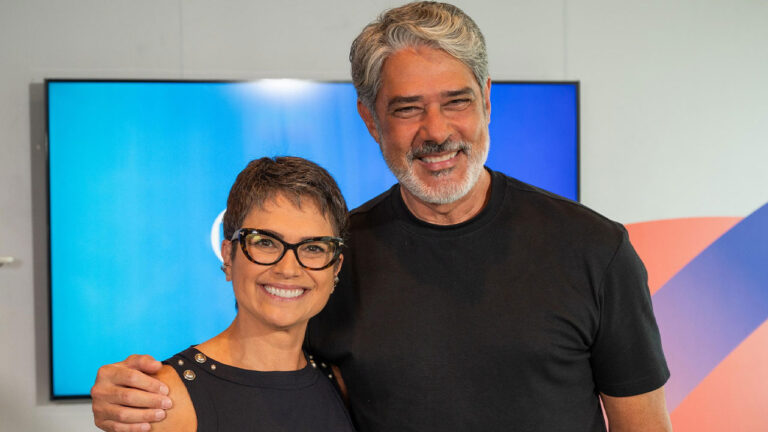 Jornalistas Sandra Annenberg e William Bonner sorriem abraçados em evento Annenberg usa óculos e blusa preta Bonner veste camiseta preta básica