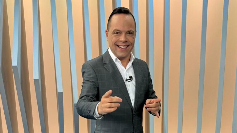 Thiago Gardinali, de terno cinza e camisa branca, sorri para a câmera em estúdio com painel de listras verticais em tons claros ao fundo, apontando com as duas mãos em gesto descontraído.