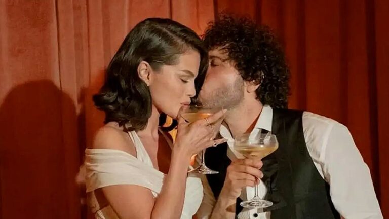 Homem e mulher vestidos de forma elegante aparecem diante de cortina vermelha, cada um segurando uma taça de coquetel. Eles se inclinam um em direção ao outro em momento íntimo, com expressão de romance e cumplicidade, sugerindo cena sofisticada ambientada em festa ou evento noturno.