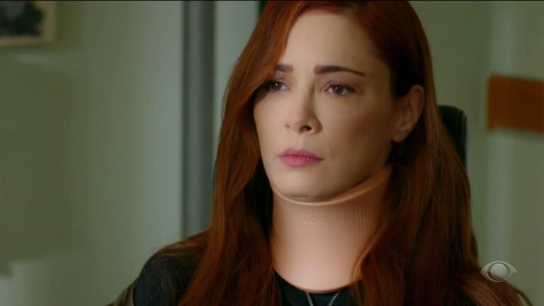 Seniz, uma mulher ruiva de cabelos longos ruivos, aparece em close, com expressão séria, usando colar cervical bege e blusa escura; ela está em ambiente interno com fundo desfocado, em uma cena de Cruel Istambul