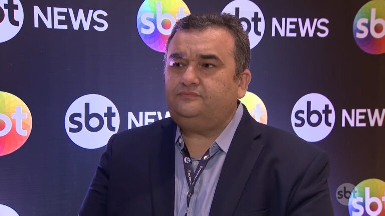 Sérgio Bringel, CEO do Grupo Norte de Comunicação, vestindo um terno escuro e camisa azul aparece em frente a um painel com logotipos do SBT News. Ele usa crachá e tem expressão séria, indicando participação em evento relacionado à emissora