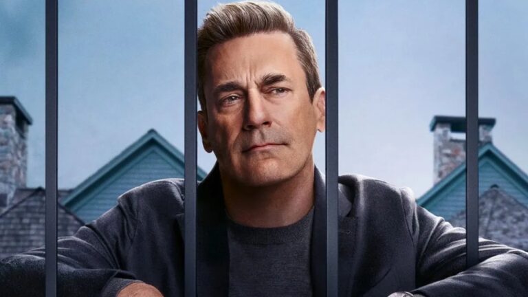 Homem de expressão séria atrás de grades em frente a casas com telhados verdes em cena de série de drama