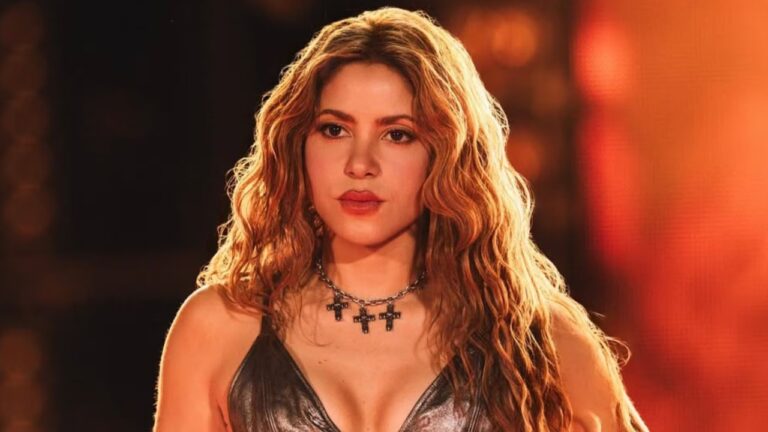 Mulher com cabelos longos e ondulados posa sob iluminação quente de palco, usando top escuro e colar com pingentes de cruz, com expressão séria diante de fundo alaranjado desfocado.