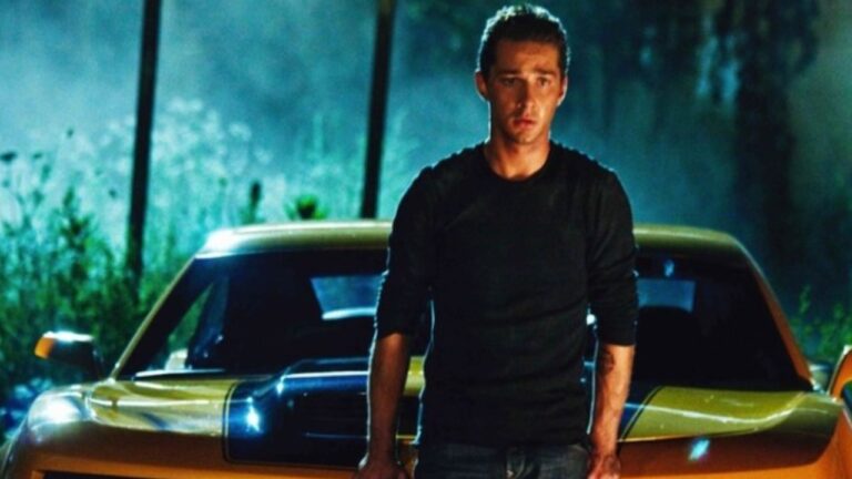 Homem jovem, vestindo camiseta preta e jeans, está em pé diante de um carro esportivo amarelo com faixa preta no capô, durante a noite. A iluminação azulada e úmida ao fundo sugere ambiente externo após chuva. Ele encara a frente com expressão tensa e determinada, enquanto os faróis e o brilho da lataria destacam o clima dramático da cena.