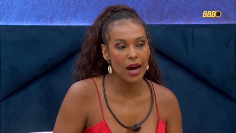 Mulher de vestido vermelho e cabelo preso em rabo de cavalo conversa dentro da casa do BBB, usando colar com microfone; logotipo do programa aparece no canto superior direito.