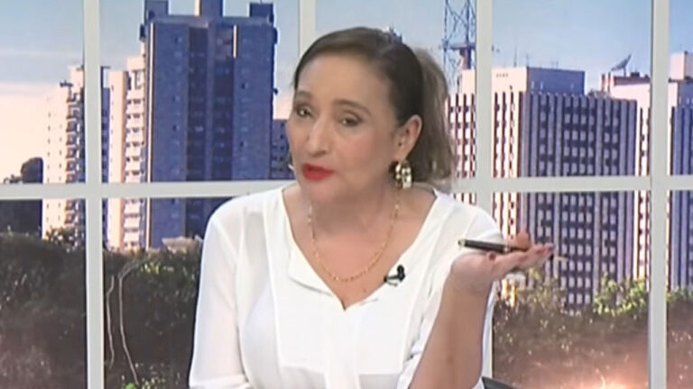 Sonia Abrão de blusa branca, batom vermelho e brincos dourados, gesticulando com uma caneta na mão no cenário do programa A Tarde é Sua com prédios ao fundo
