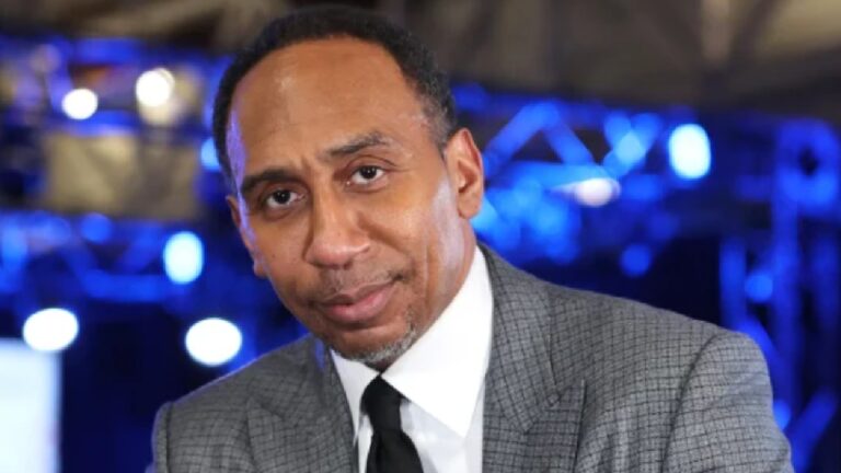 Homem de terno cinza e gravata preta olha diretamente para a câmera, em ambiente com iluminação azul e estruturas metálicas desfocadas ao fundo, sugerindo estúdio ou evento.