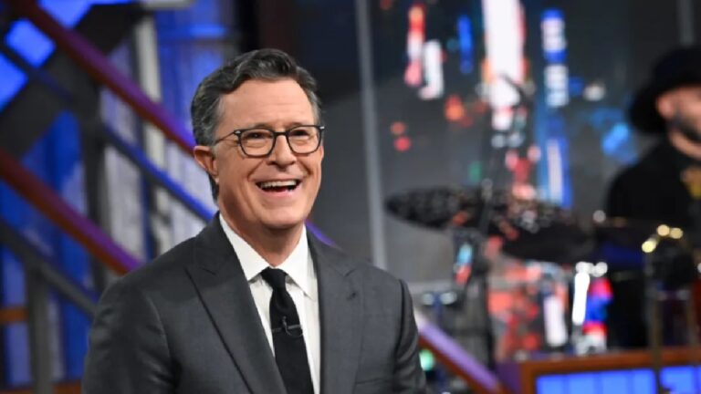 Apresentador de TV veste terno escuro, camisa branca e gravata preta enquanto sorri durante gravação em estúdio. Ele usa óculos de armação fina e está sentado diante de cenário iluminado com tons de azul e vermelho. Ao fundo, é possível ver parte da banda e equipamentos musicais, indicando ambiente de talk show ou programa de entretenimento noturno.