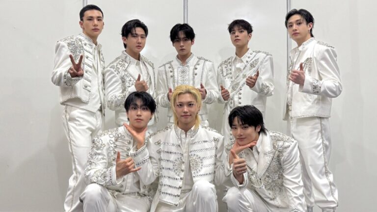 Grupo de k-pop posa nos bastidores usando figurinos brancos com detalhes prateados antes de apresentação em evento musical