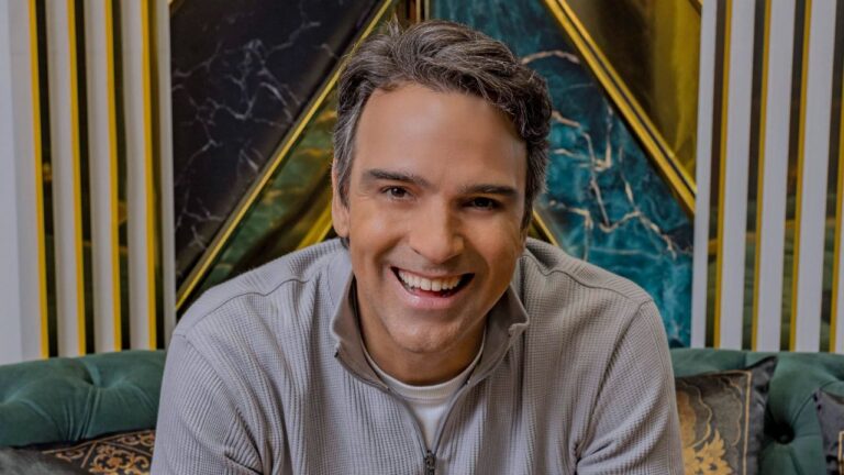 Apresentador Tadeu Schmidt sorri para a câmera vestindo blusa de zíper cinza, sentado em sofá verde com painéis geométricos de mármore ao fundo