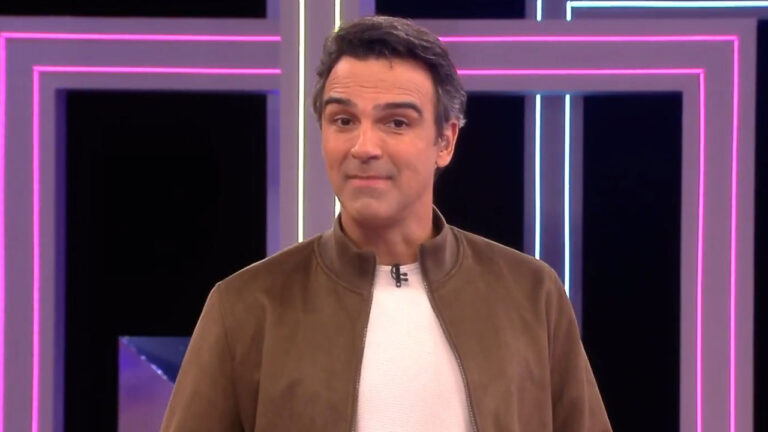 Tadeu Schmidt encara a câmera usando jaqueta marrom e camiseta branca, com cenário de luzes neon roxas ao fundo, durante apresentação do Big Brother Brasil na Globo