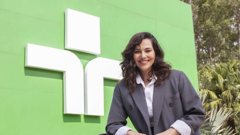 Mulher sorridente usa blazer cinza e camisa branca, posando em frente ao logotipo verde e branco da TV Cultura em ambiente externo com árvores ao fundo.
