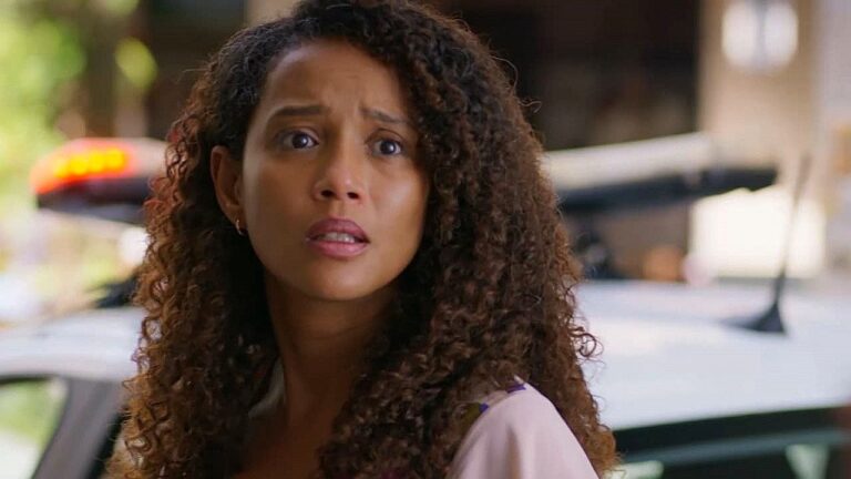 Mulher de cabelos cacheados aparece em primeiro plano com expressão de surpresa e preocupação, olhando para algo fora de quadro. Ao fundo, parcialmente desfocado, há uma viatura policial com luzes de emergência acesas, sugerindo situação tensa em ambiente externo urbano.