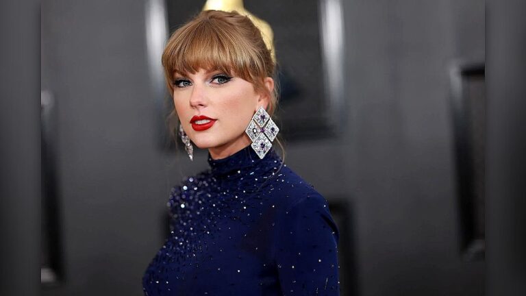 Mulher loira posa para fotos em evento de tapete vermelho, usando vestido azul-marinho de gola alta com aplicações brilhantes. Ela tem franja reta, cabelo preso em coque e maquiagem marcante com batom vermelho. Os brincos grandes e geométricos chamam atenção, enquanto o fundo desfocado sugere ambiente de premiação ou cerimônia formal.