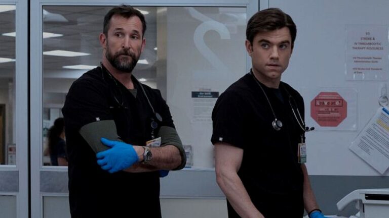 Dois profissionais de saúde vestindo uniformes médicos pretos e com estetoscópios no pescoço estão em um corredor de hospital. Um deles usa luvas azuis e mantém os braços cruzados, enquanto ambos olham para o lado com expressão séria, diante de portas de vidro e avisos fixados na parede, sugerindo momento de tensão ou atenção a uma situação clínica.