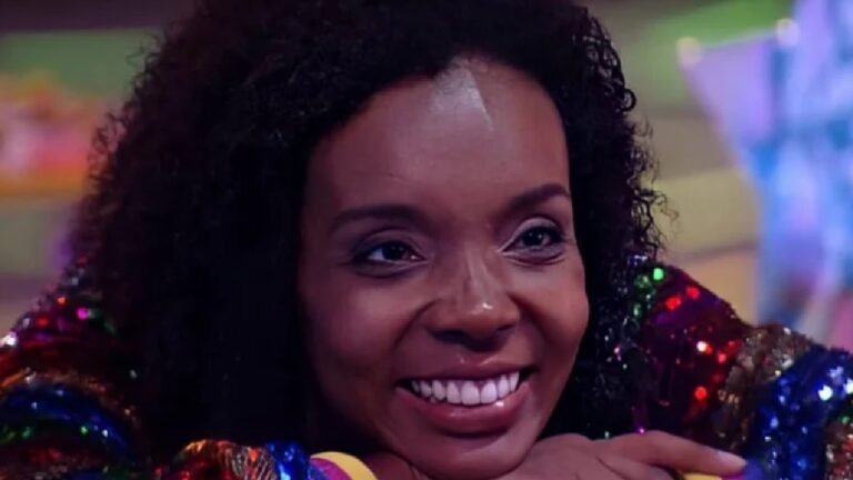 Mulher negra com cabelos crespos aparece em close, sorrindo amplamente e apoiando o queixo sobre as mãos. Ela veste roupa com mangas coloridas e brilhantes, em tons de vermelho, verde e azul. O ambiente ao fundo tem iluminação vibrante e desfocada, sugerindo cenário de programa de TV ou reality show.