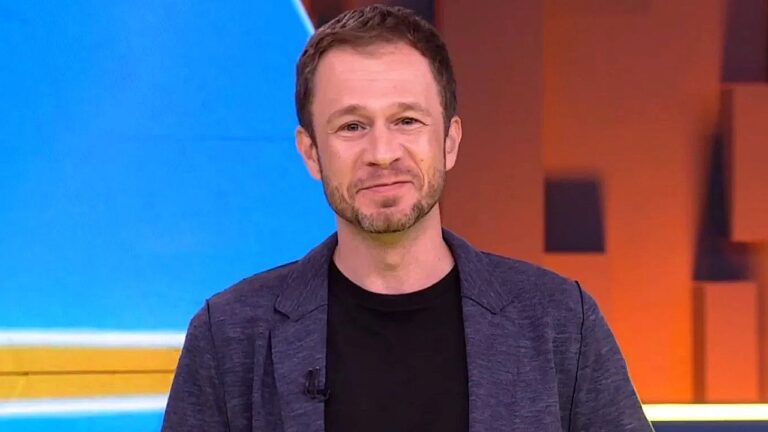 Homem sorri levemente durante participação em programa de televisão, vestindo camiseta preta e blazer azul. Ele está em estúdio com fundo colorido em tons de azul e laranja, iluminado por luzes de palco.
