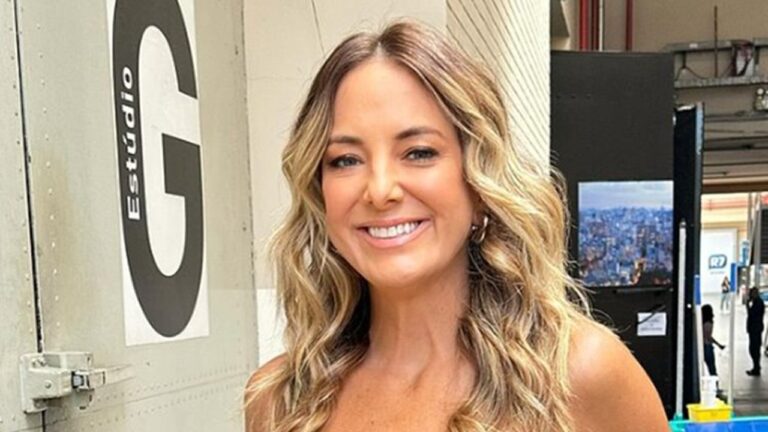 Apresentadora de cabelo loiro cacheado sorri em corredor de estúdio de TV com placa Estúdio G ao fundo