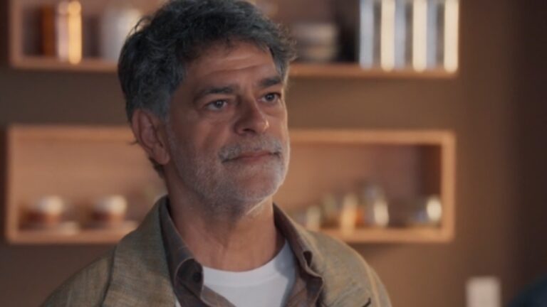 Homem de meia-idade aparece em um ambiente interno com iluminação quente e aconchegante. Ele tem cabelos grisalhos, barba por fazer e expressão serena, olhando levemente para a frente. Usa uma camiseta branca por baixo de uma camisa marrom e um casaco em tom neutro. Ao fundo, há prateleiras embutidas com objetos decorativos e iluminação suave, criando um clima acolhedor e sofisticado.