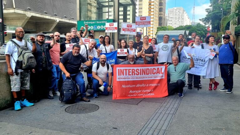 Grupo numeroso de homens e mulheres adultos reunidos em calçada urbana participa de manifestação em frente a prédios altos, muitos com o punho direito erguido em gesto de protesto, vestindo roupas casuais como camisetas, calças jeans, mochilas e bonés, alguns usando óculos. No centro do grupo há uma faixa vermelha com o texto “INTERSINDICAL – instrumento de luta e organização da classe trabalhadora – Nenhum direito a menos – Avançar rumo a novas conquistas”. Pessoas ao redor seguram cartazes e faixas menores com logotipos e textos como “TVT” e “Jornalistas”