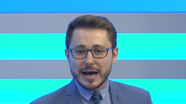 Apresentador de telejornal com óculos, terno e gravata fala em estúdio com fundo azul durante edição de noticiário ao vivo