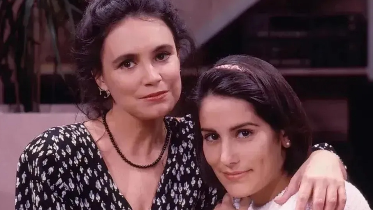 Duas mulheres aparecem abraçadas em um ambiente interno. A mais velha, com cabelo escuro preso e blusa preta estampada, envolve a mais jovem com o braço sobre os ombros. A jovem, de cabelo curto escuro e faixa na cabeça, apoia-se nela e olha para a câmera com leve sorriso, transmitindo proximidade e afeto.