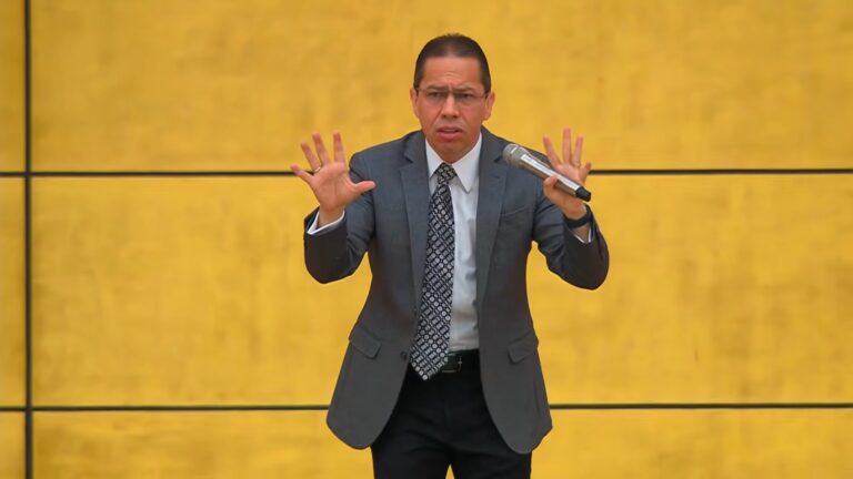 Jadson Santos, homem de terno cinza e gravata segura um microfone e gesticula com as duas mãos erguidas enquanto fala em um palco com fundo amarelo liso, indicando participação na Sessão do Descarrego, exibida pela Record.