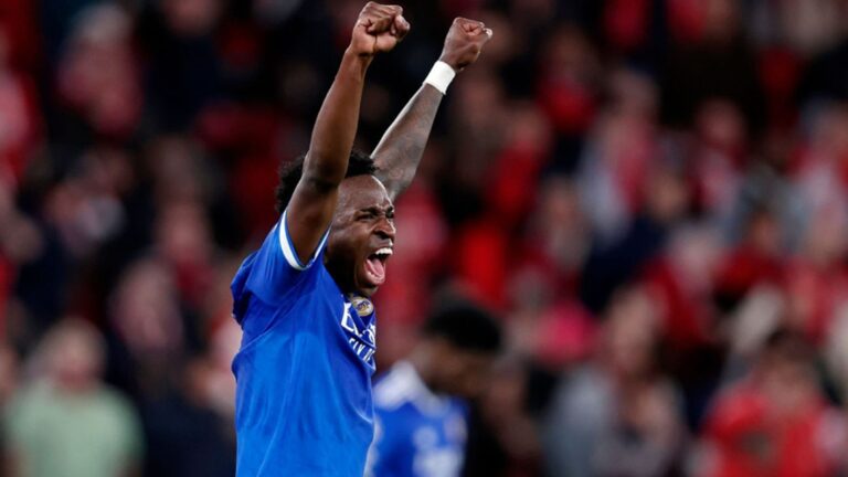 Vini Jr., jogador de futebol com uniforme azul do Real Madrid, comemora em campo com os dois braços erguidos e punhos cerrados, gritando em direção à torcida; ao fundo, arquibancadas desfocadas repletas de espectadores vestidos predominantemente de vermelho, cor característica do Benfica.