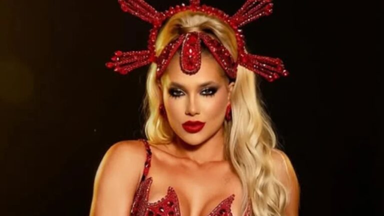 Mulher loira posa em estúdio com fundo escuro, usando figurino de espetáculo em tom vermelho intenso composto por sutiã estruturado e adornado com pedrarias brilhantes, decote profundo realçando o colo e alças finas, maquiagem marcante com olhos fortemente delineados em preto, cílios volumosos, sobrancelhas definidas e batom vermelho escuro, expressão confiante e sensual com olhar direto para a câmera, cabelos longos loiros ondulados caindo sobre os ombros