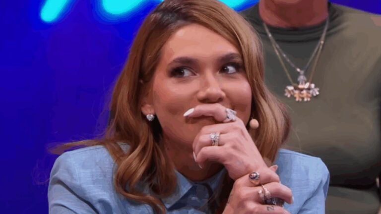 Mulher com cabelos longos e ondulados, usando blazer azul-claro, aparece em close durante um programa de TV. Ela leva a mão à boca, exibindo vários anéis e unhas longas, com expressão entre surpresa e emoção. Ao fundo, luzes em tons de azul e roxo sugerem estúdio de gravação, e um microfone discreto está preso ao rosto, indicando participação ao vivo.