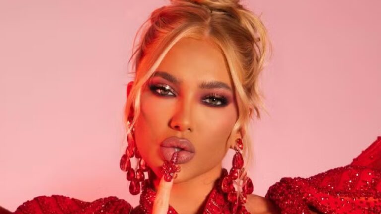 Mulher loira com maquiagem marcante e roupa vermelha faz gesto de silêncio com dedo nos lábios em ensaio fotográfico promocional