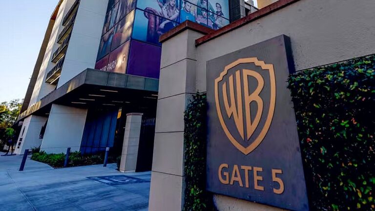 Fachada de prédio com o logotipo da Warner Bros. e a inscrição “WB Gate 5” em destaque em uma placa na entrada. O edifício moderno, com painéis de vidro e cartazes promocionais na parte superior, sugere acesso a estúdio ou complexo de produção audiovisual.