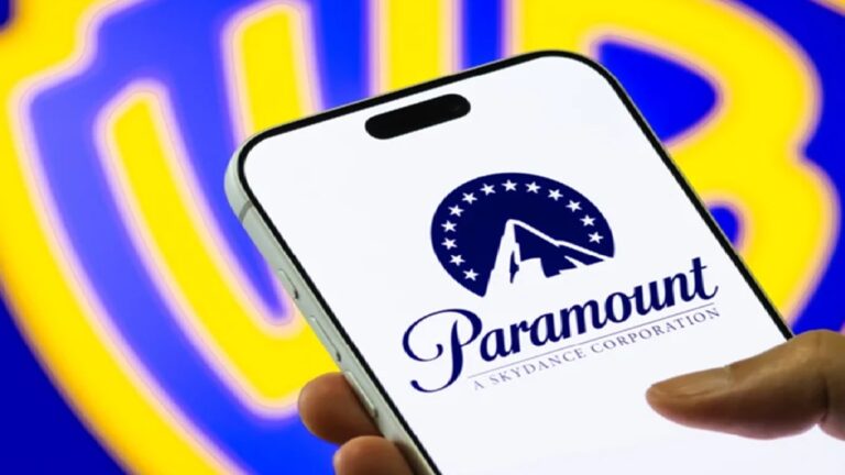 Smartphone exibe na tela o logotipo da Paramount, com o nome da empresa e o símbolo da montanha cercada por estrelas. Ao fundo, desfocado, aparece o escudo azul e dourado da Warner Bros., sugerindo contexto de mercado audiovisual ou possível parceria entre estúdios de entretenimento.