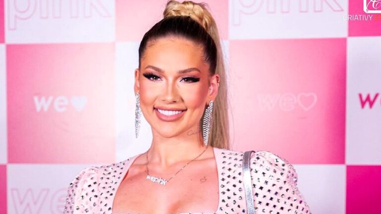 Mulher posa sorrindo para fotos em evento, diante de painel quadriculado em tons de rosa com logotipos ao fundo. Ela usa vestido claro com aplicações brilhantes, brincos longos e cabelo preso em rabo de cavalo alto, compondo visual glamouroso de tapete vermelho.